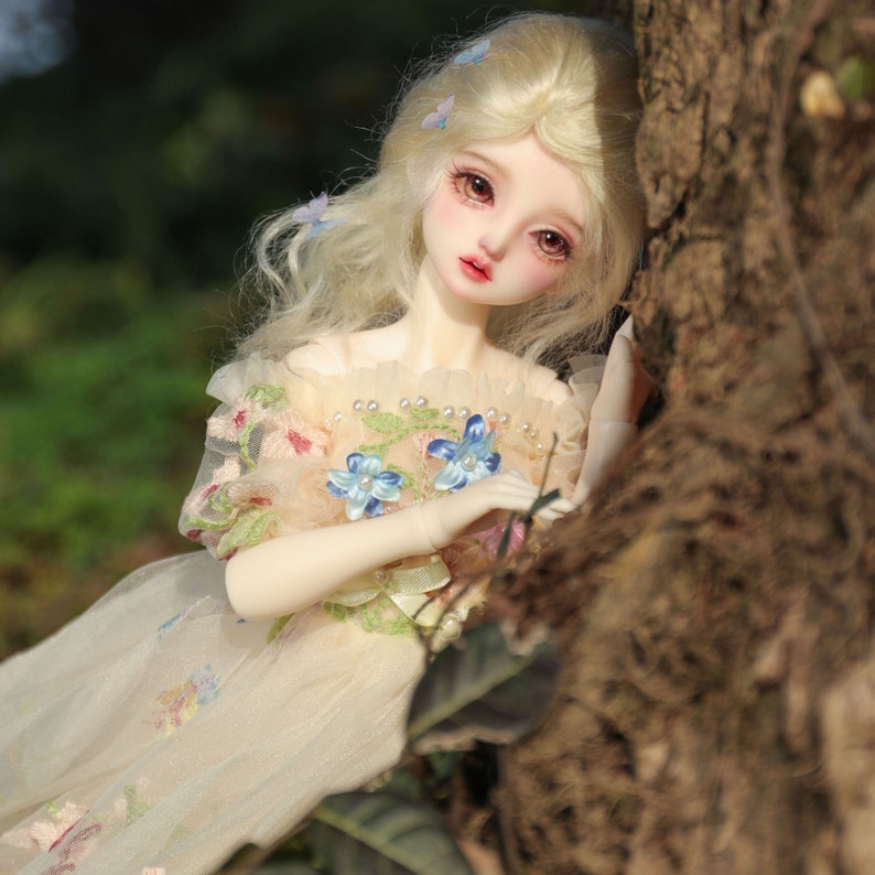 May include: A porcelain doll with blonde hair and blue eyes, wearing a white dress with floral embroidery and blue butterfly accents. The doll is leaning against a tree trunk.