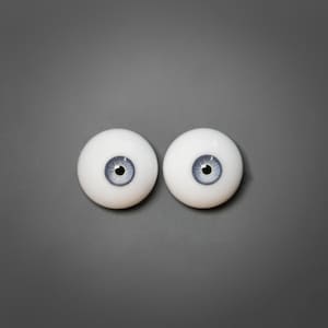 May include: Two white doll eyes with light blue irises and black pupils are displayed against a gray background. The eyes are round and have a realistic appearance, suitable for doll making or craft projects.