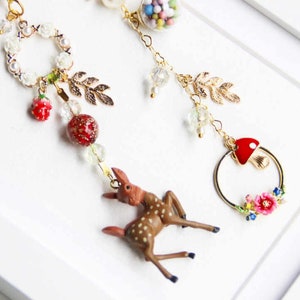 Puede incluir: Un collar de cadena dorada con una variedad de dijes, que incluyen una fresa roja y blanca, un hongo rojo y dorado, una pequeña figura de ciervo y una pequeña corona con perlas blancas. El collar también incluye un pequeño frasco de vidrio lleno de caramelos coloridos.