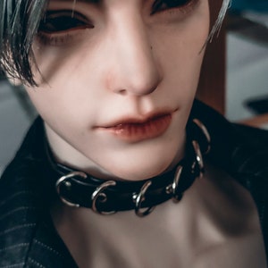 May include: A close-up of a person wearing a black leather choker with silver rings. The choker is worn around the neck and has a simple design.