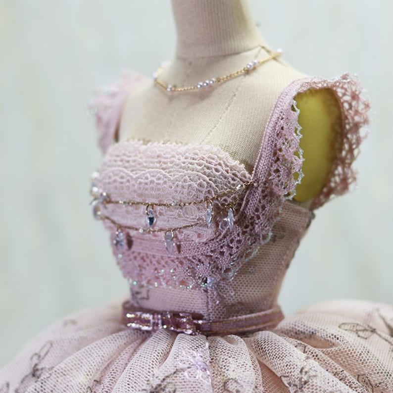 Op de afbeelding: Een roze tule- en kantjurk met een delicate parelketting en een gouden ketting met kristallen accenten. De jurk heeft een getailleerd lijfje en een volle rok.