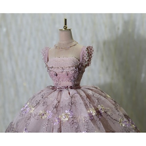 Op de afbeelding: Een roze tule baljurk met bloemenborduursel en een parelversierd lijfje. De jurk staat op een mannequin met een gouden standaard.