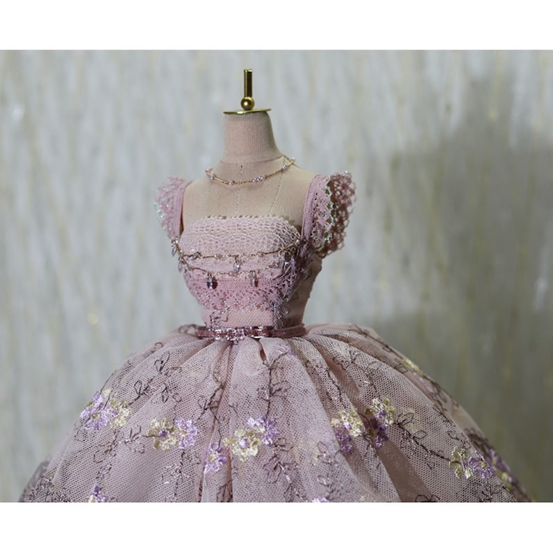 Op de afbeelding: Een roze tule baljurk met bloemenborduursel en een parelversierd lijfje. De jurk staat op een mannequin met een gouden standaard.