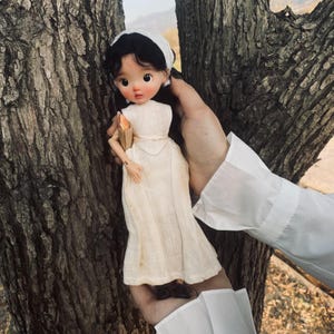 May include: A vintage-style doll with black hair, rosy cheeks, and large eyes, wearing a cream-colored dress and a white headscarf. The doll is holding a small, tan-colored object. The doll is being held in front of a tree trunk.
