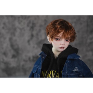 May include: A doll with short, brown hair wearing a black hoodie with the word "NIRVANA" printed on it and a blue denim jacket.