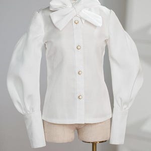 May include: A white blouse featuring a large bow at the neck and long, puffed sleeves. The blouse has a button-down front with gold-coloured buttons. The cuffs are long and buttoned. The blouse is displayed on a mannequin.