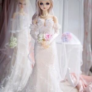 May include: Two dolls wearing white wedding dresses with lace details and veils. One doll is holding a bouquet of white and pink flowers.