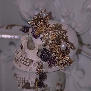 Può includere: Un teschio bianco con una corona floreale dorata e viola. La corona è composta da foglie, bacche e perle.