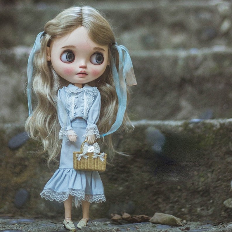 May include: A vintage-style doll with large eyes and long, wavy blonde hair styled in pigtails with blue ribbons. The doll wears a light blue dress with lace trim and a matching skirt. She holds a small woven basket with a white bow.