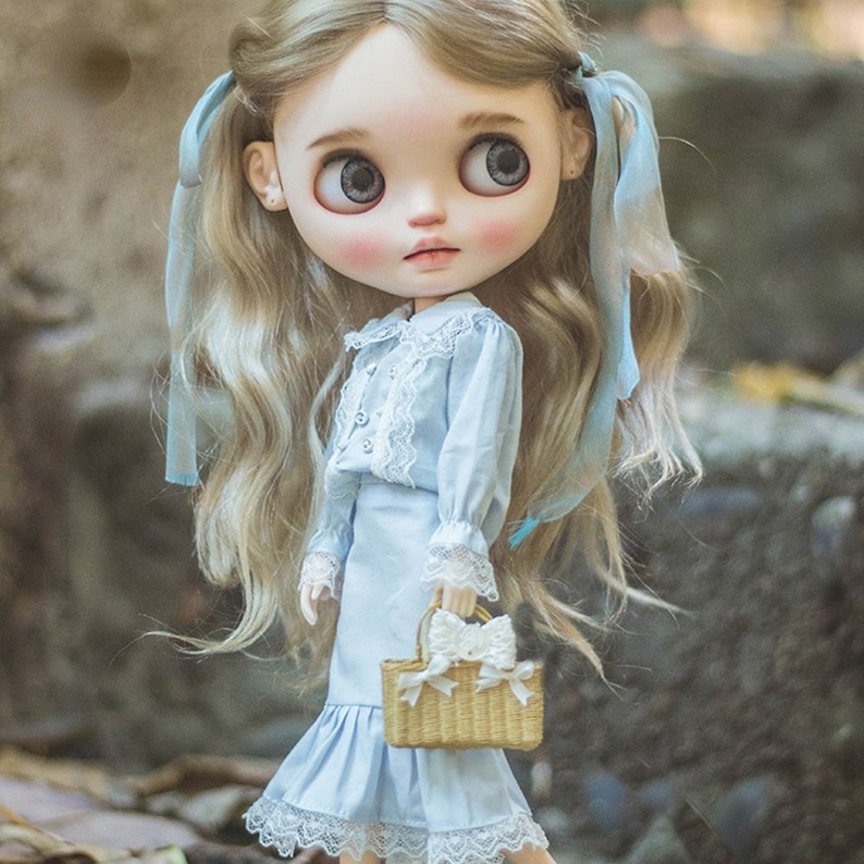 May include: A Blythe doll with long, wavy blonde hair styled with light blue ribbons. The doll wears a light blue dress with white lace trim and a matching skirt. She holds a small, woven basket with a white bow.