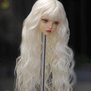 Può includere: Testa di bambola con pelle chiara, occhi blu e labbra rosse. La bambola ha lunghi capelli biondo platino ondulati con frangia. La testa è montata su un supporto in metallo argentato, con uno sfondo sfocato.