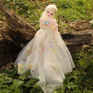 May include: A porcelain doll with blonde hair wearing a white tulle dress with floral embroidery. The doll is sitting on a tree trunk in a grassy area.