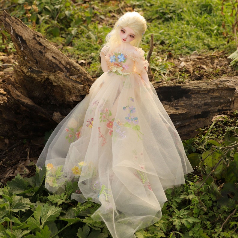May include: A porcelain doll with blonde hair wearing a white tulle dress with floral embroidery. The doll is sitting on a tree trunk in a grassy area.