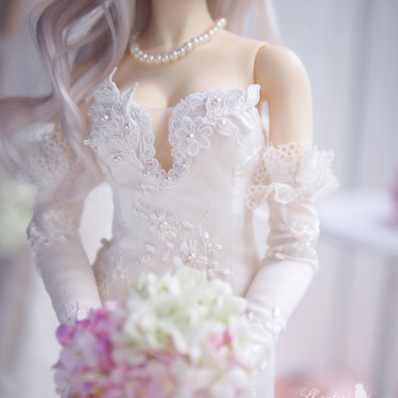 May include: A white bridal gown with a lace bodice and long sleeves. The gown has a sweetheart neckline and is adorned with pearl embellishments. The bride is holding a bouquet of white and pink flowers.