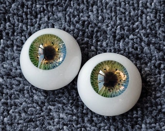 Puede incluir: Dos ojos de muñeca realistas con iris verdes, azules y marrones. Los ojos están colocados sobre un fondo gris difuso.