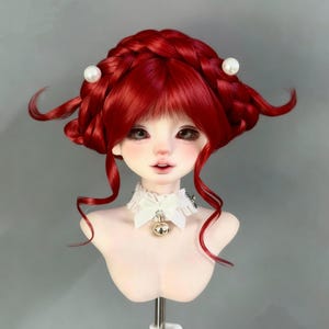 May include: A doll head with a vibrant red wig, styled with a braided crown and fringe. The doll has a pale complexion, detailed facial features, and a white lace collar with a gold bell pendant. Two white pearl hair accessories are visible.