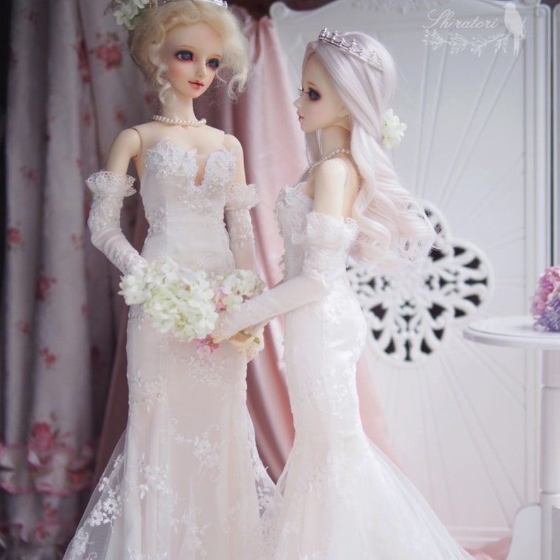 May include: Two dolls wearing white wedding dresses with lace details and veils. The doll on the left is holding a bouquet of white flowers. The doll on the right has a silver tiara.