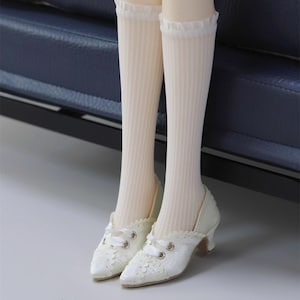 May include: A pair of cream-colored, lace-covered heels with a small heel and ribbon laces. The shoes are paired with cream-colored, ribbed knee-high socks with a ruffled trim.