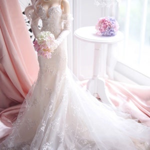 May include: A white wedding dress with lace detailing and a long train. The dress is being held by a doll with a bouquet of pink and white flowers.
