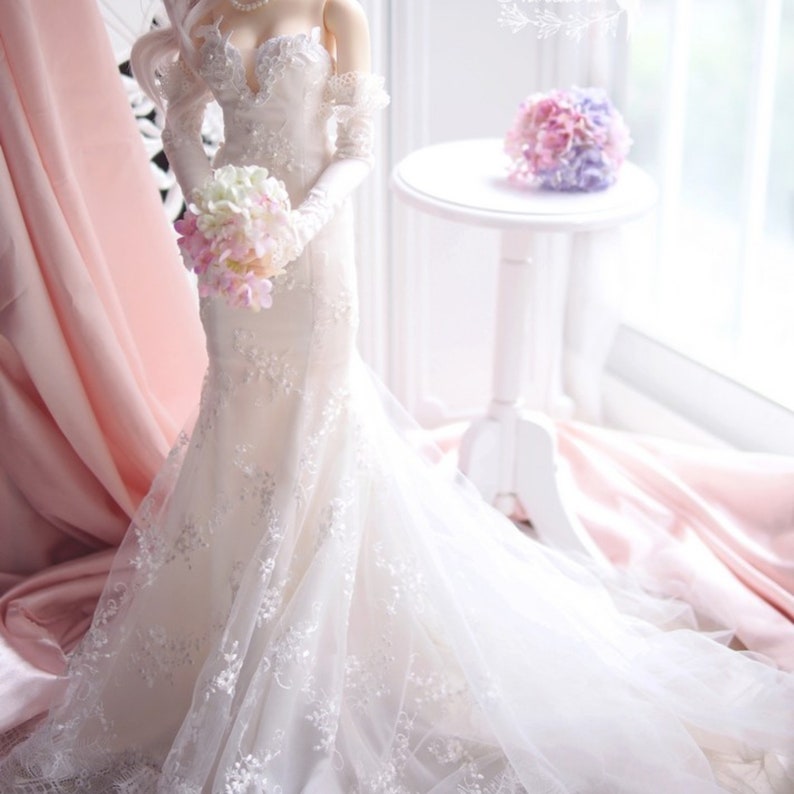 May include: A white wedding dress with lace detailing and a long train. The dress is being held by a doll with a bouquet of pink and white flowers.