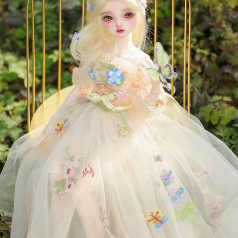 May include: A porcelain doll wearing a white tulle dress with floral embroidery and a green butterfly on her hand. The doll has blonde hair and blue eyes.