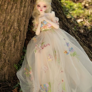 May include: A porcelain doll with blonde hair wearing a white tulle dress with floral embroidery. The doll is leaning against a tree trunk.