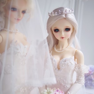 May include: Two porcelain dolls wearing white wedding dresses. The doll on the right is wearing a silver crown and a pearl necklace. Both dolls are wearing veils.