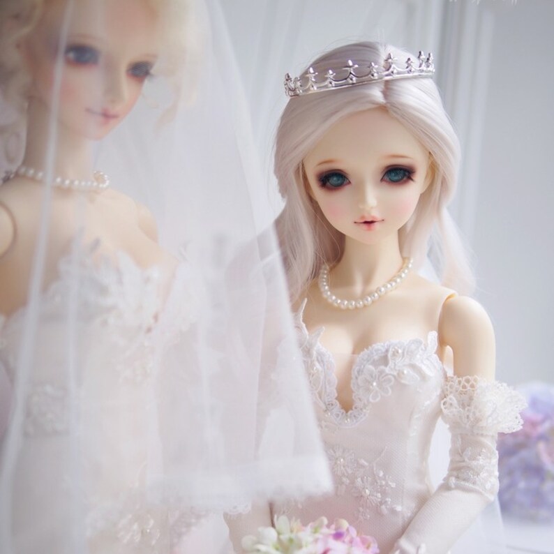 May include: Two porcelain dolls wearing white wedding dresses. The doll on the right is wearing a silver crown and a pearl necklace. Both dolls are wearing veils.