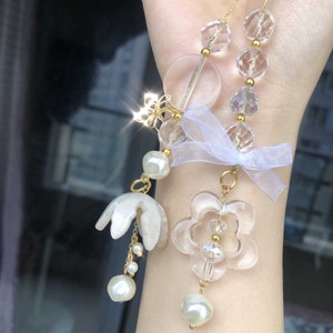 May include: A delicate necklace with clear crystal beads, gold accents, and a white flower charm. The necklace also features a white pearl charm and a white ribbon.