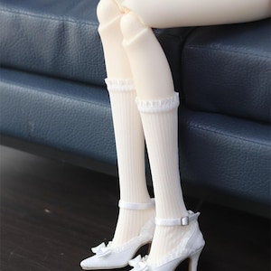 Op de afbeelding: Poppenbenen in gebroken witte geribde kniekousen met ruches en enkelbandjes. De pop draagt witte pumps met strikken en enkelbandjes. De achtergrond is een donkere houten vloer en een blauwe leren bank.