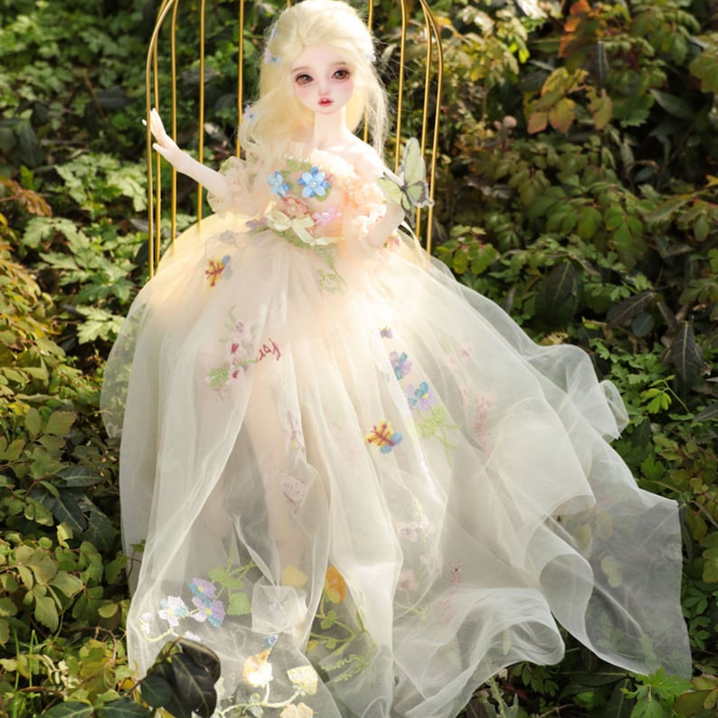 May include: A porcelain doll with long blonde hair wearing a white dress with floral embroidery and a butterfly on the shoulder. The doll is sitting on a gold birdcage.