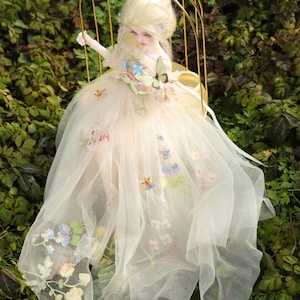 May include: A porcelain doll wearing a white tulle dress with floral embroidery and a butterfly on the bodice. The doll has blonde hair and blue eyes.