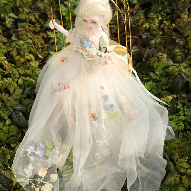 May include: A porcelain doll wearing a white tulle dress with floral embroidery and a butterfly on the bodice. The doll has blonde hair and blue eyes.