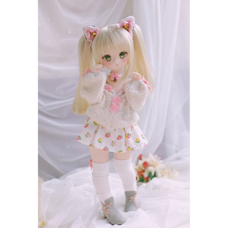 May include: A doll with blonde hair wearing a white fuzzy sweater with pink bows, a white skirt with pink flowers, white knee-high socks, and grey shoes. The doll is wearing a pink cat ear headband.