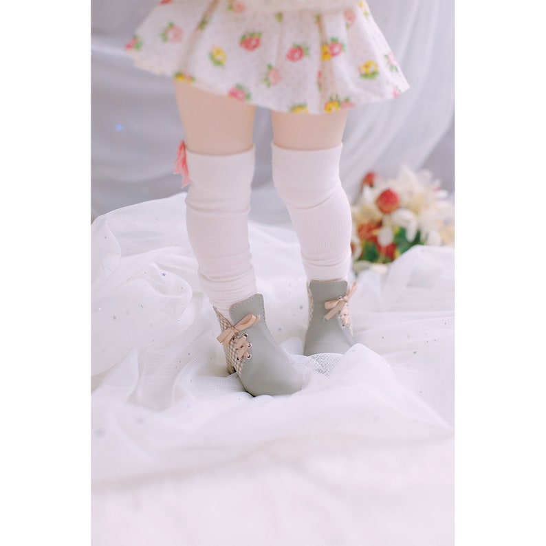 May include: A pair of gray ankle boots with a lace-up front and a pink bow detail. The boots are worn by a child wearing a white skirt with a floral pattern and white knee-high socks.