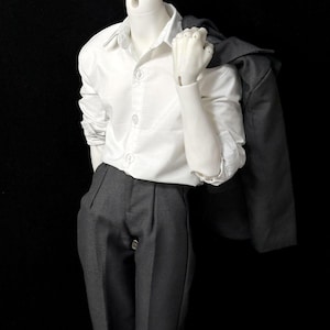 Puede incluir: Una camisa blanca abotonada, pantalones grises oscuros y una blazer gris oscuro sobre el hombro. El conjunto se exhibe en un maniquí sobre un fondo negro. La camisa tiene mangas largas y está abotonada.