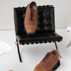 Puede incluir: Una silla de cuero marr&oacute;n con un asiento de cuero negro y patas cromadas. Dos zapatos de cuero marr&oacute;n est&aacute;n en la silla y en el suelo.