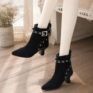 May include: A pair of black ankle boots featuring a pointed toe, a low kitten heel, and a decorative strap with a silver-toned buckle. The boots are accented with silver metal eyelets along the strap and a row of small silver studs on the side panel.