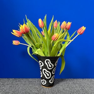Op de afbeelding: Een boeket gele en rode tulpen in een zwart-wit gepatroneerde vaas. De vaas heeft een wit abstract ontwerp op een zwarte achtergrond.