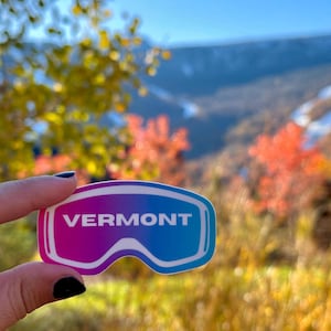 May include: A sticker in the shape of ski goggles with the word "VERMONT" in white. The sticker has a gradient of pink and blue. The background is a blurred view of a mountain and fall foliage.