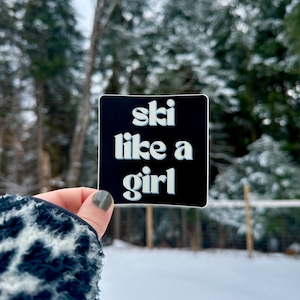 May include: A black and white sticker with the text "ski like a girl" in a retro font.