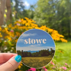 May include: A round sticker with a blue sky, green trees, and yellow flowers. The sticker says "stowe vermont" in white text.