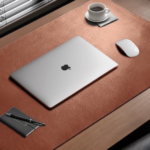 Peut inclure: Un sous-main rectangulaire en cuir marron avec des bords cousus. Le tapis comprend un ordinateur portable argenté avec un logo Apple, une souris d'ordinateur blanche, un petit carnet noir avec un stylo noir et une tasse à café blanche sur soucoupe.