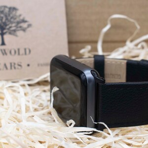 May include: A black smartwatch with a textured black leather strap and a black buckle. The watch face is dark and reflective. In the background, a tan card displays a tree logo and the text COTSWOLD WEARS, surrounded by light-colored wood shavings.