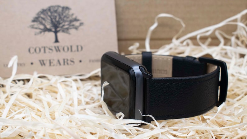 May include: A black smartwatch with a textured black leather strap and a black buckle. The watch face is dark and reflective. In the background, a tan card displays a tree logo and the text COTSWOLD WEARS, surrounded by light-colored wood shavings.
