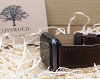 May include: A brown leather watch band with a silver Apple Watch. The band is stitched with brown thread and has a buckle closure. The watch is sitting on a bed of shredded paper.