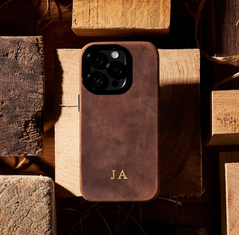 K&ouml;nnte beinhalten: Eine braune Leder Handyh&uuml;lle mit einem Kamera-Ausschnitt und den Initialen "JA" in Gold gepr&auml;gt.
