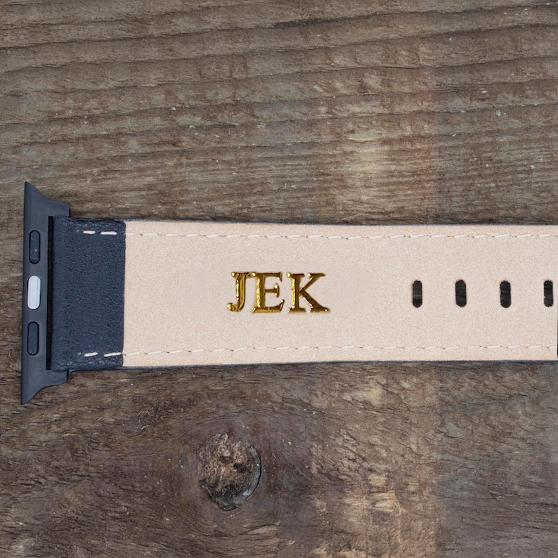 May include: A watch strap featuring a dark blue exterior and a light beige interior lining. The interior surface is embossed with the gold-colored letters JEK. The strap includes a black metal connector piece and a series of adjustment holes.