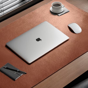 May include: A brown leather desk mat with a personalised name "Olivia" in black lettering. A silver laptop, a black pen, a small black notebook, a white mouse, and a white coffee cup are on the mat.