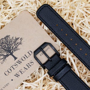 May include: Black leather watch strap with a black buckle, next to a tan tag with the text "Cotswold Wears" and a tree graphic. The strap is textured and has several holes for adjustment.